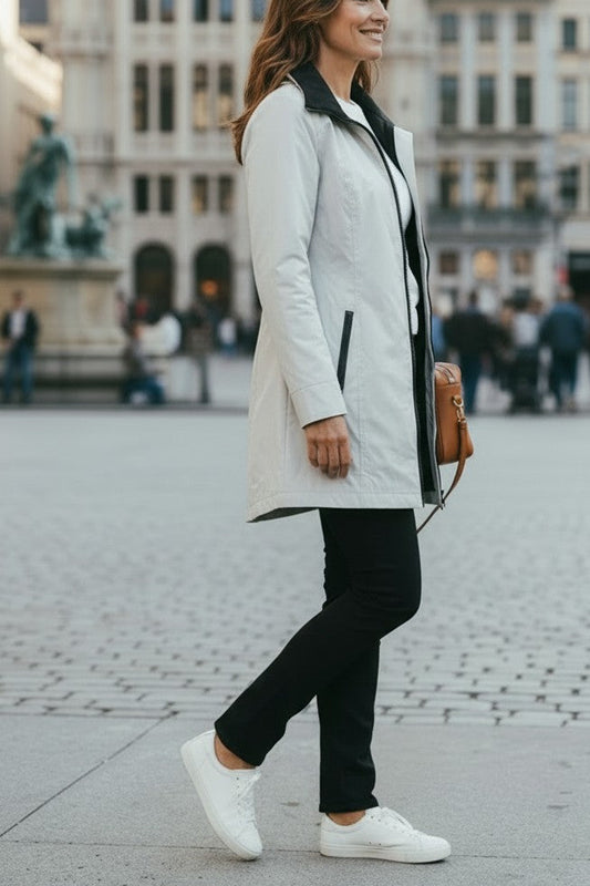 A woman walking in a city square wearing a light gray coat, black pants, white sneakers, and carrying a brown shoulder bag, with historic buildings blurred in the background.