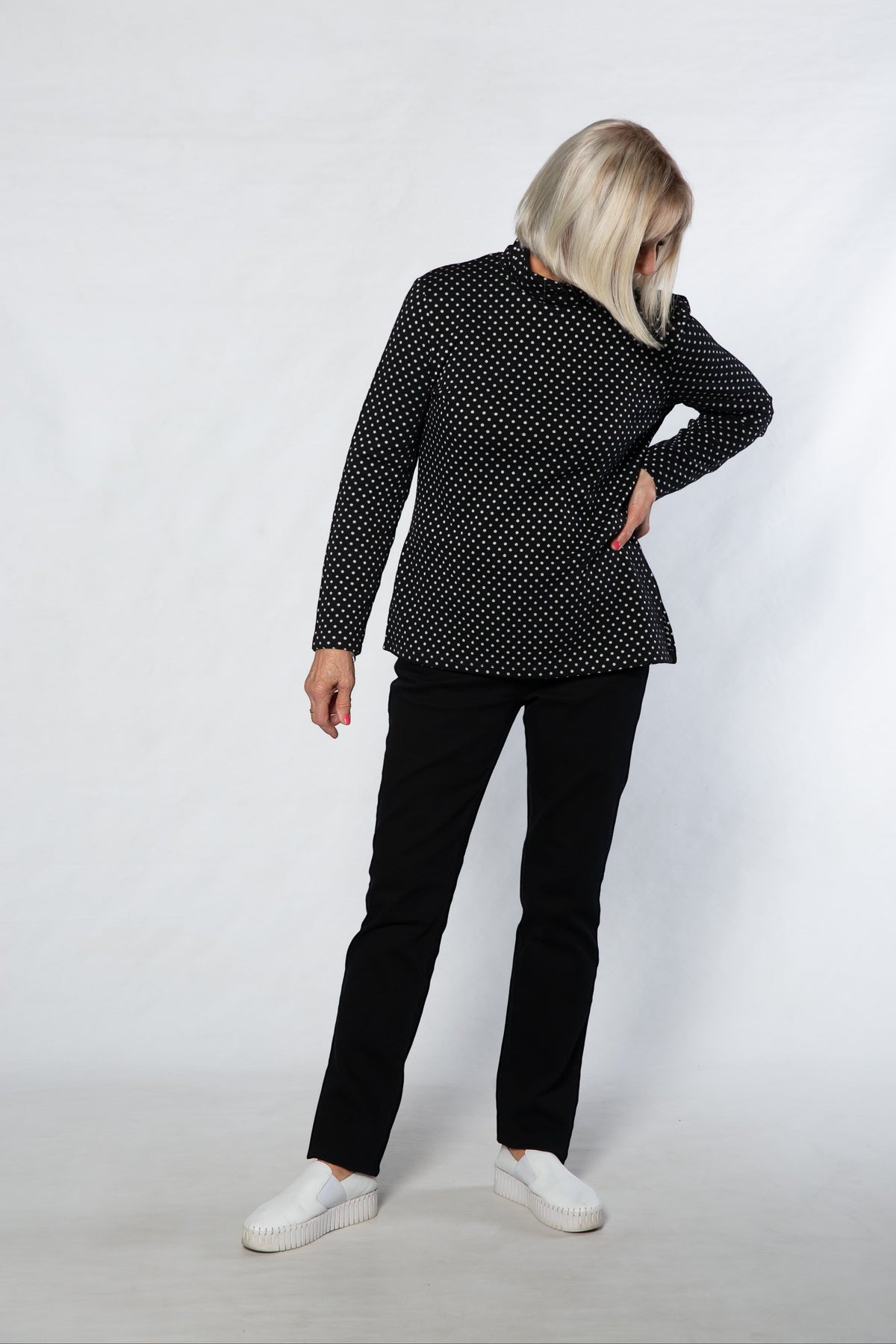 Equinox Clothing black and white polka dot mock turtle skivvy worn by a woman with black trousers and white sneakers.
