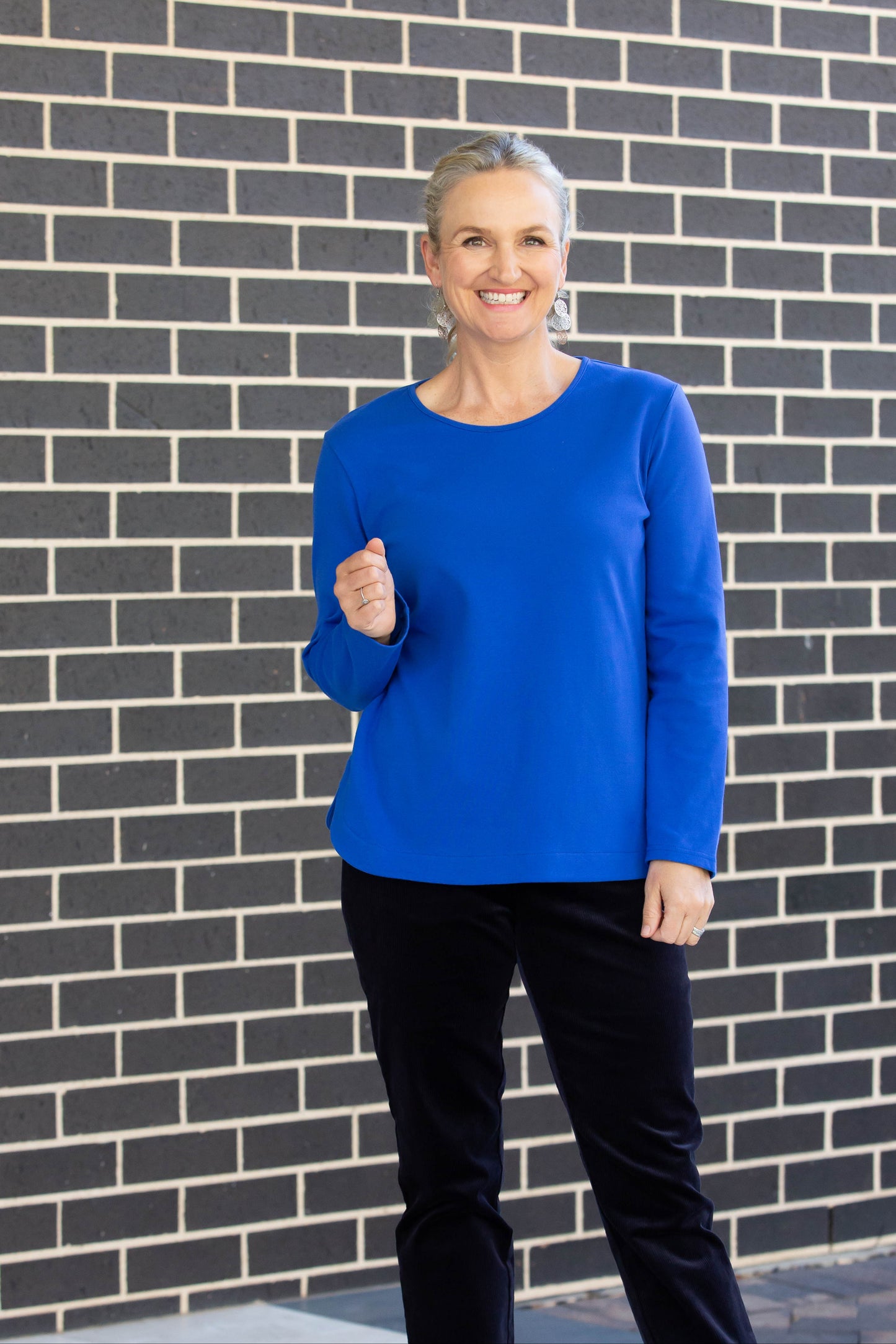 A model wearing a long sleeve stretch cotton round neck top in blue color with a crew neckline and a curved hemline, paired with black pants and black shoes.