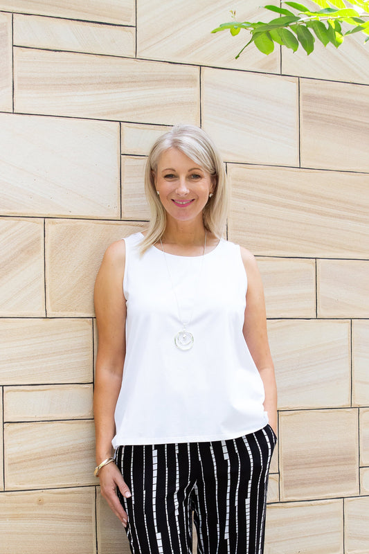 A woman wearing a sleeveless white stretch cotton top and striped pants, standing against a brick wall background.