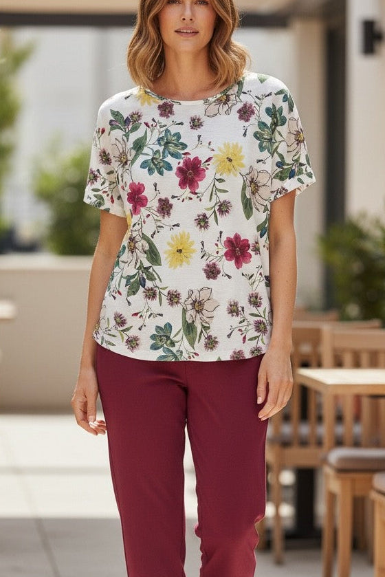Woman wearing a floral blouse and burgundy pants walking outdoors near tables and chairs.
