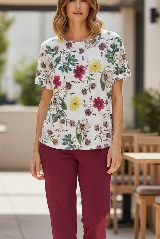 Woman wearing a floral blouse and burgundy pants walking outdoors near tables and chairs.