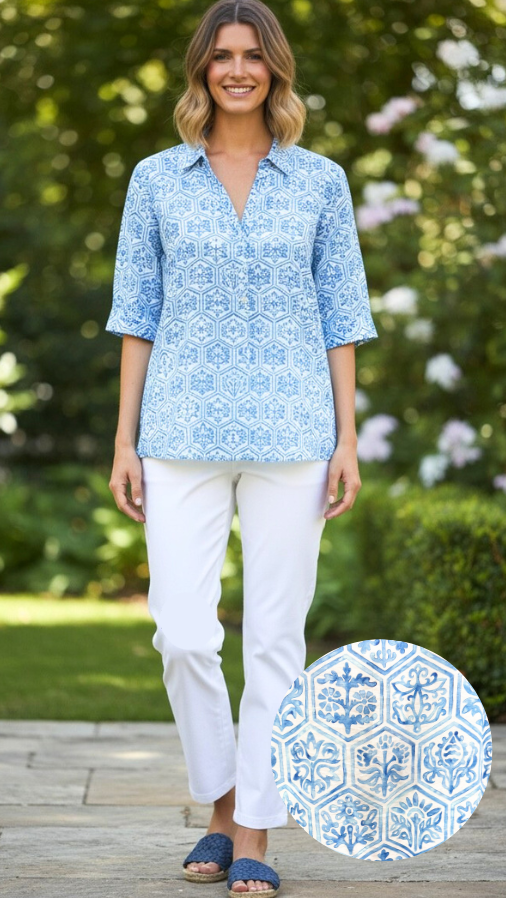 Woman wearing a blue patterned shirt and white pants standing outdoors with greenery in the background