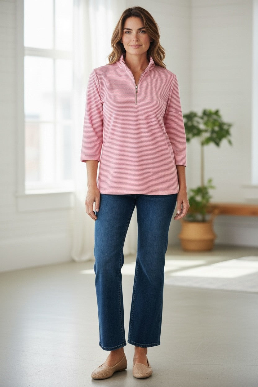 Woman wearing a pink striped shirt and blue pants standing in a bright room with a plant in the background.
