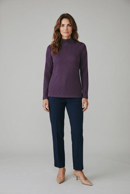 A woman modeling a long-sleeve mock neck knit top with a purple and white micro-dot pattern, paired with navy blue trousers and beige heels, against a light gray studio background.