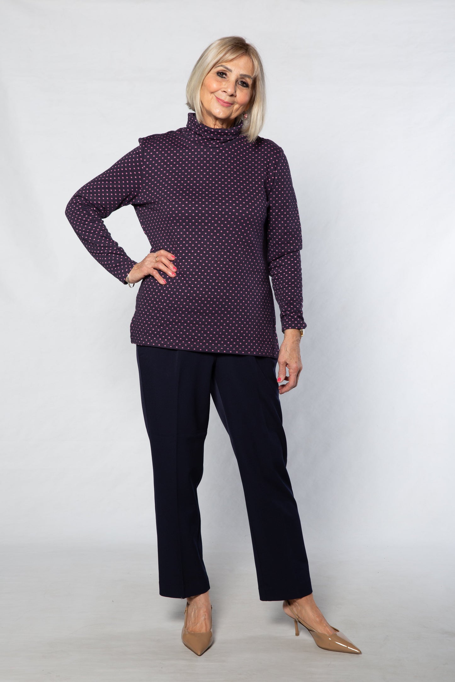 Woman wearing a purple polka dot top and black pants against a white background