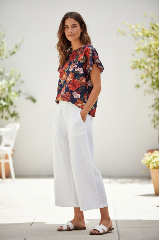 Woman wearing an Equinox Clothing floral top and white pants standing outdoors with plants in the background