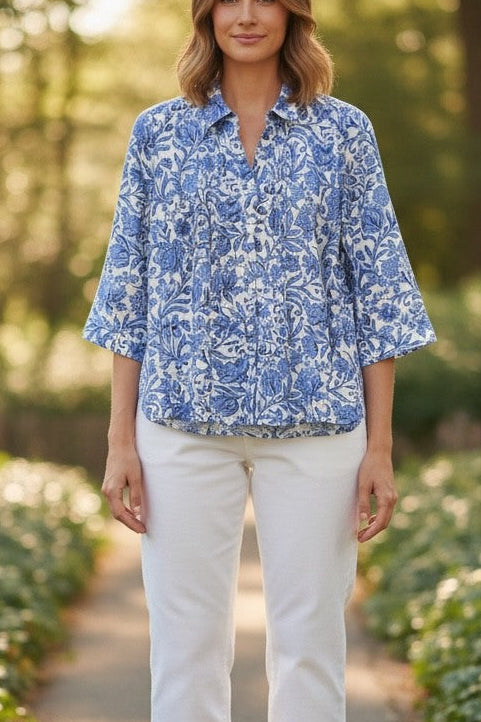 Woman in a blue floral blouse and white pants standing on a path in a garden.