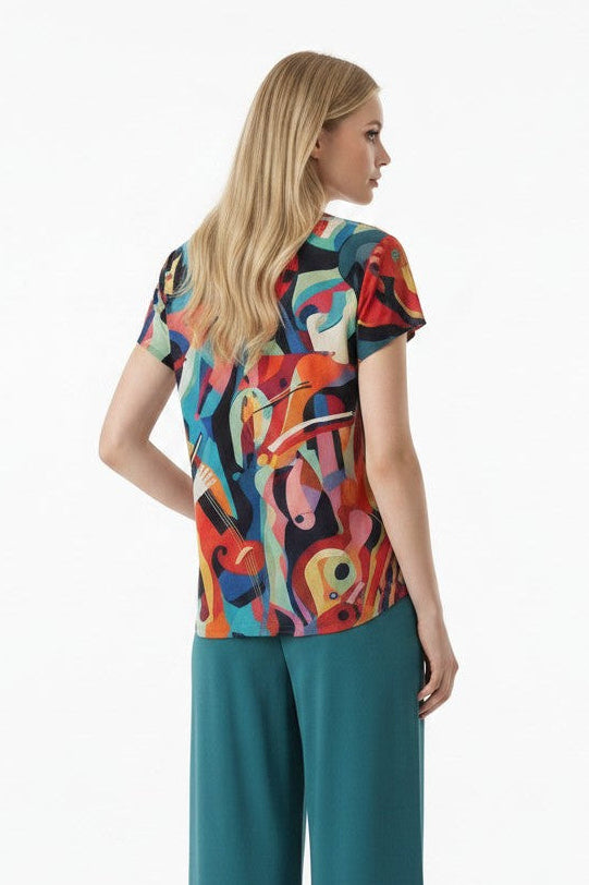 A woman stands with her back to the camera wearing a colorful abstract-patterned short-sleeve top and teal wide-leg cropped pants against a plain white studio background.