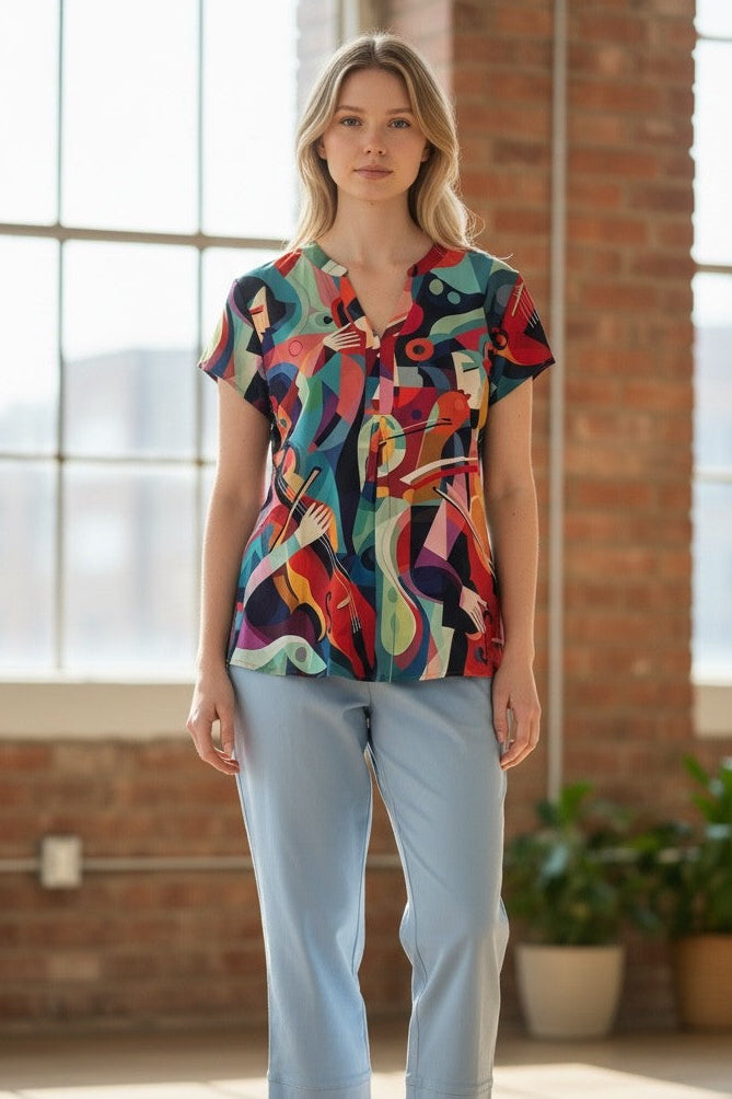 A woman stands in a bright loft-style room with brick walls and large windows, wearing a short-sleeve button-front blouse with a bold multicolor abstract print and light blue cropped pants.