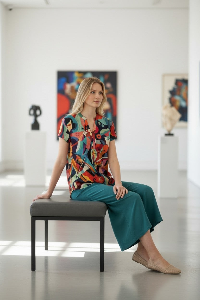 A woman sits on a gray bench in a bright, modern art gallery, wearing a colorful abstract-patterned short-sleeve blouse and teal wide-leg trousers.