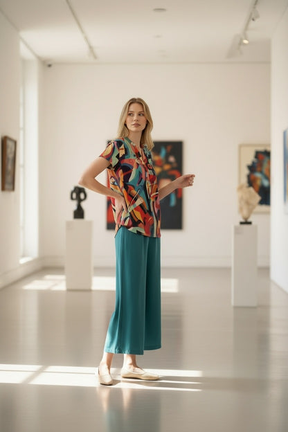 A woman stands in a bright, modern art gallery wearing a colorful abstract-patterned short-sleeve top and teal wide-leg pants. The gallery has white walls, light-colored floor, and art pieces displayed on pedestals in the background.