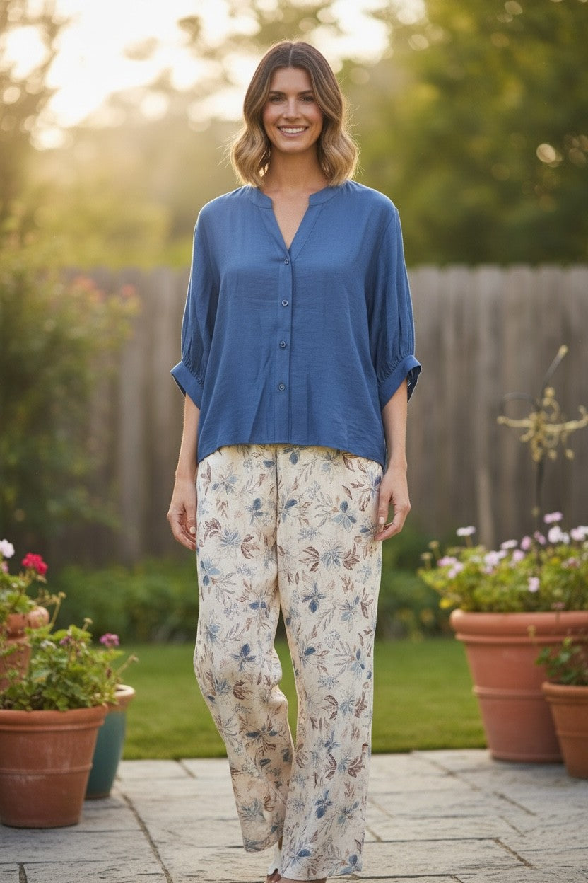 A woman stands outdoors on a stone patio wearing a blue button-front blouse with rolled sleeves and cream floral pants, with a garden and wooden fence in the background.
