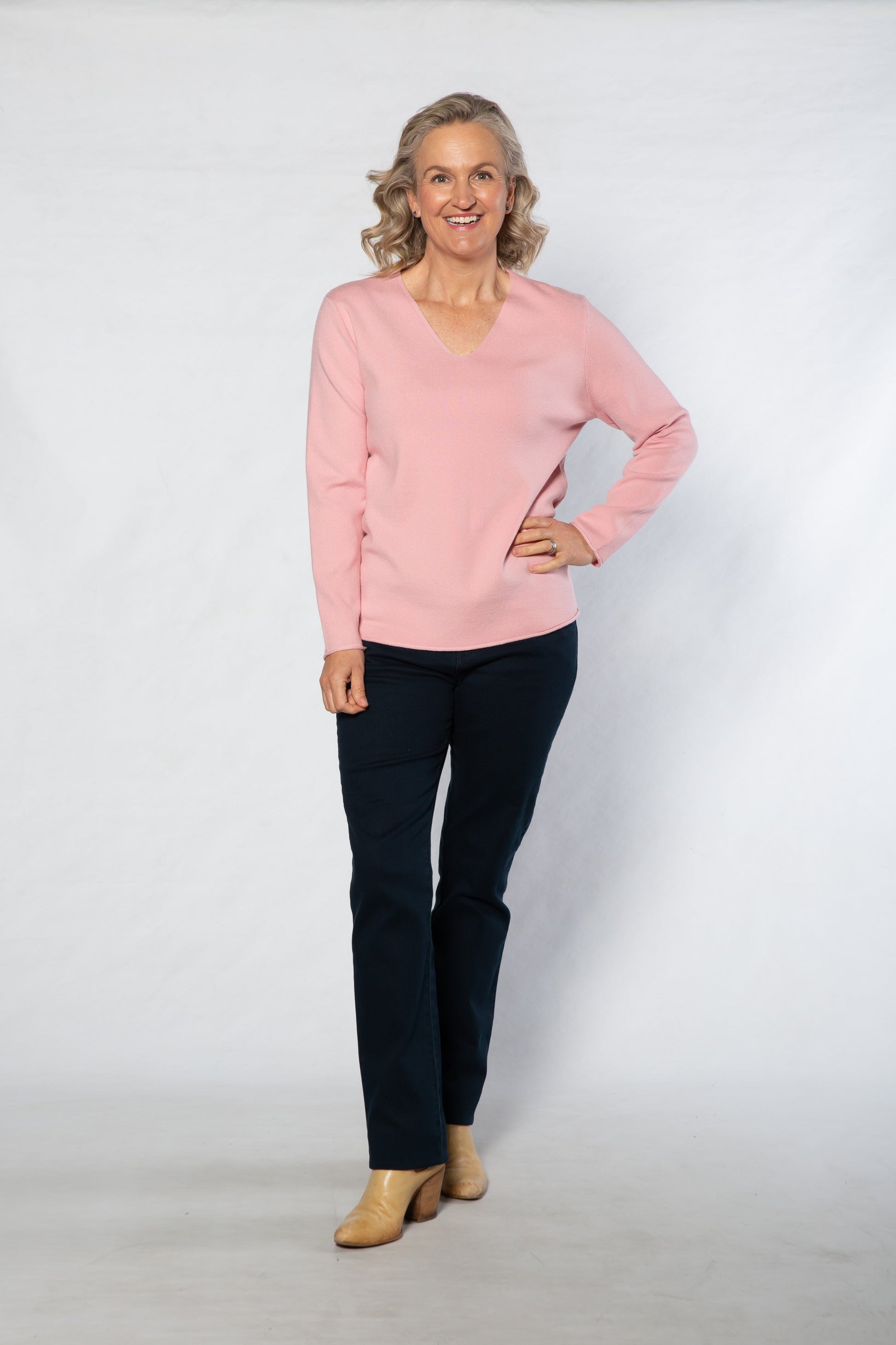 Woman wearing a pink sweater and dark pants standing against a white background