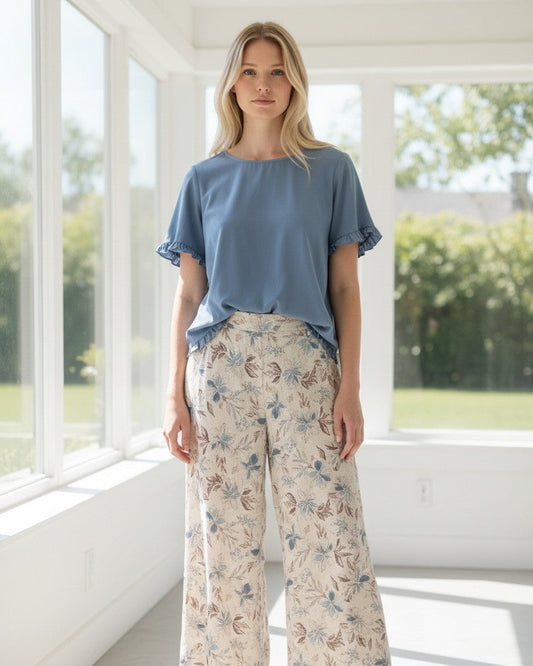 A woman stands in a bright, modern room with large windows, wearing a dusty blue short-sleeve top and beige wide-leg pants with a blue and brown floral pattern. She is wearing tan flat sandals.