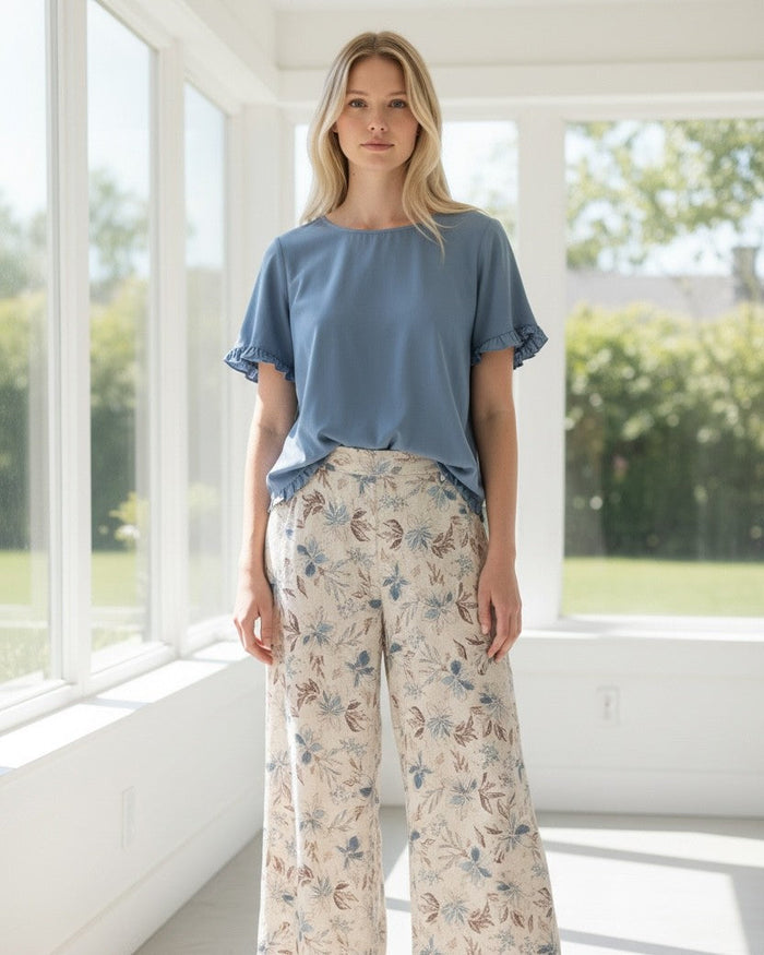 A woman stands in a bright, modern room with large windows, wearing a dusty blue short-sleeve top and beige wide-leg pants with a blue and brown floral pattern. She is wearing tan flat sandals.