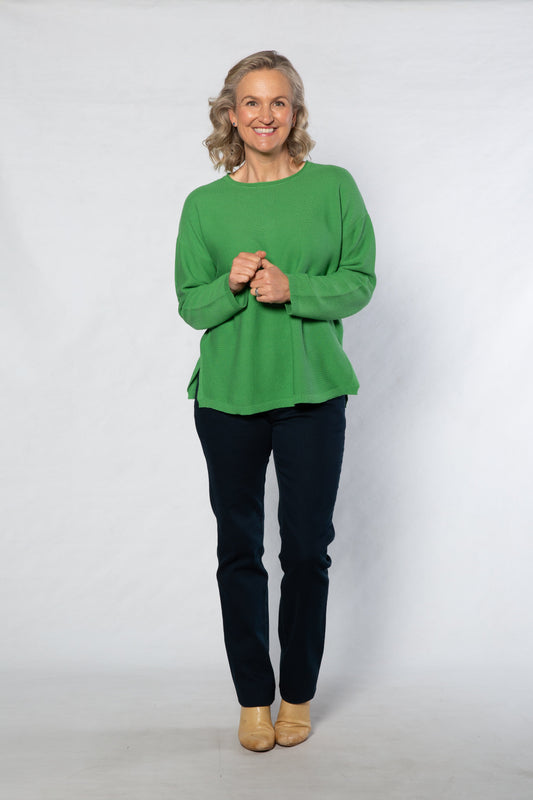 Person wearing a green sweater and dark pants standing against a white background