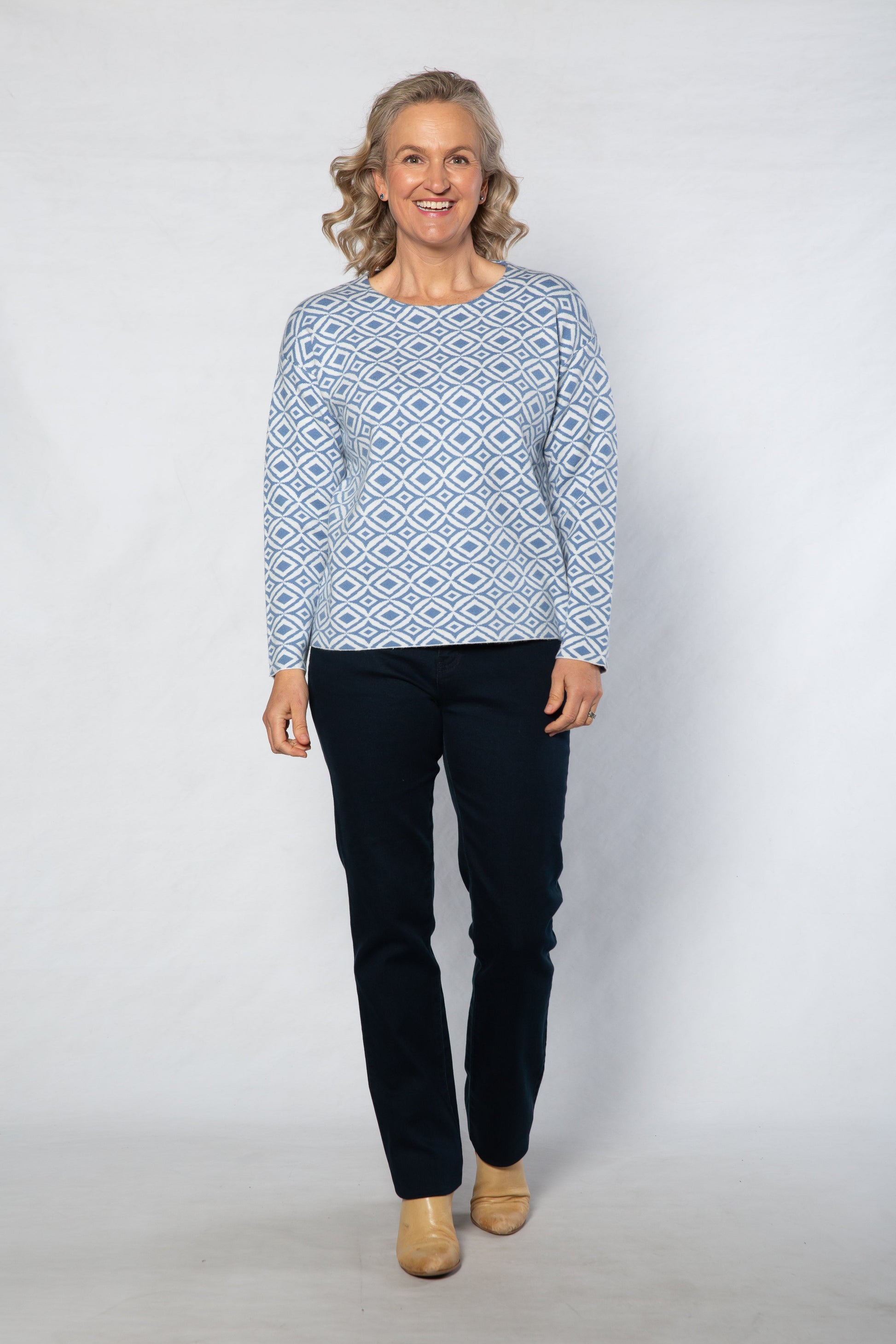 Woman wearing a patterned blue sweater and dark pants on a white background