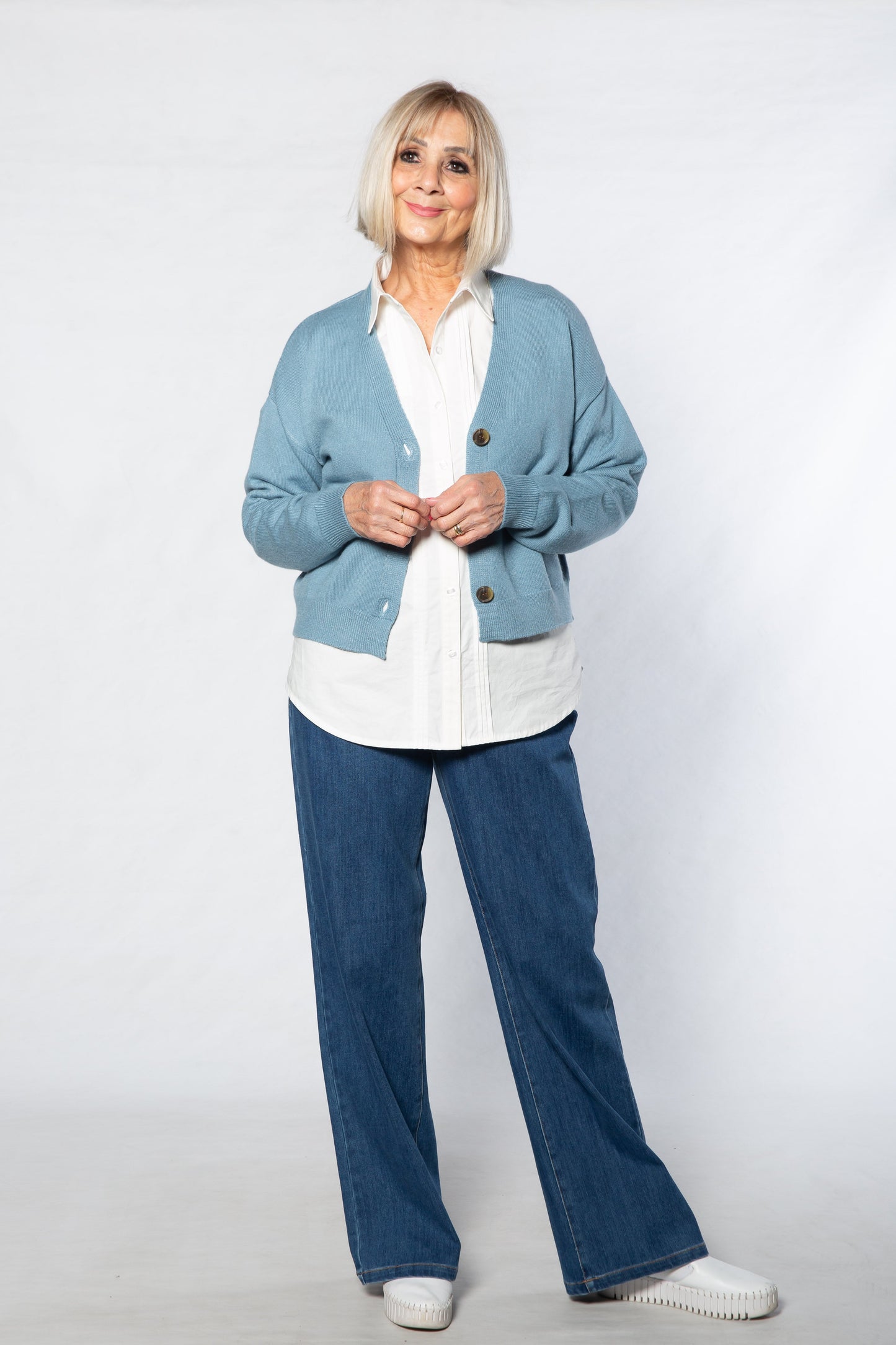 Woman wearing a light blue cardigan, white shirt, and blue jeans against a white background