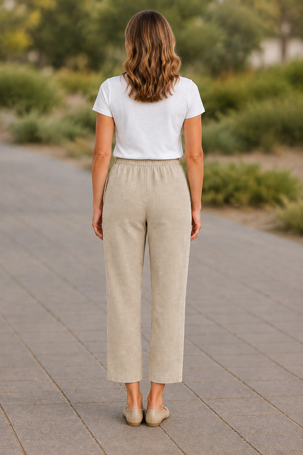 A woman viewed from behind wearing a white short-sleeve t-shirt and light beige wide-leg cropped pants, standing on a paved outdoor walkway with blurred greenery in the background.