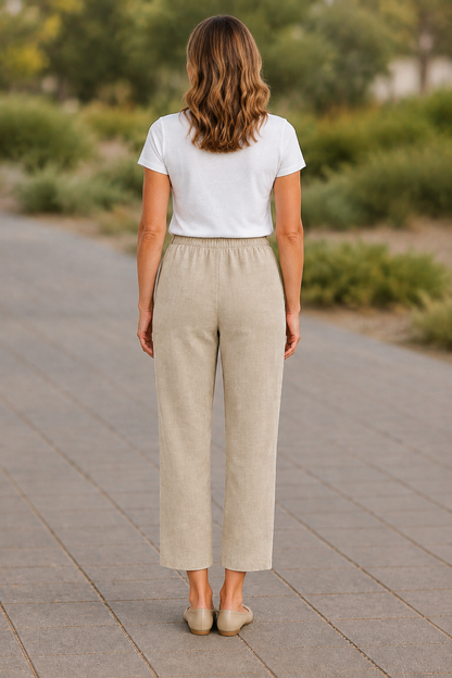 A woman viewed from behind wearing a white short-sleeve t-shirt and light beige wide-leg cropped pants, standing on a paved outdoor walkway with blurred greenery in the background.