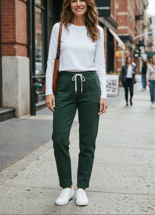 A woman stands on a city sidewalk wearing dark green drawstring pants, a white long-sleeve top, and white sneakers, with a brown shoulder bag and blurred storefronts in the background.