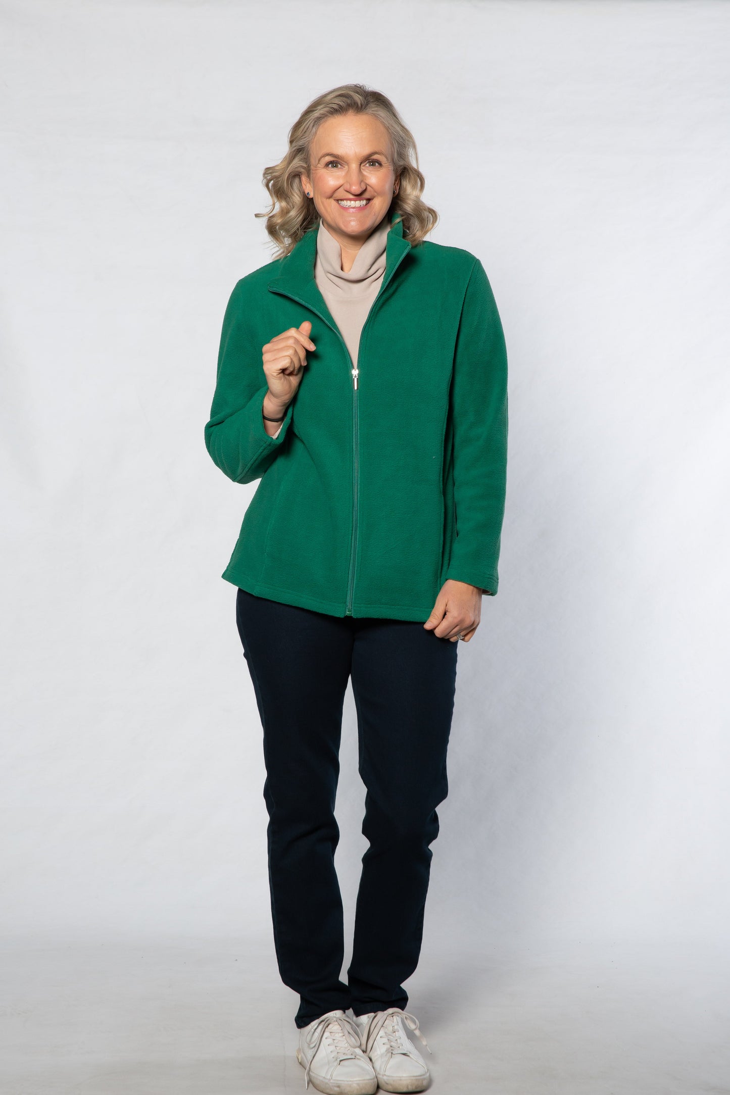 Woman wearing a green jacket, beige sweater, black pants, and white shoes on a white background