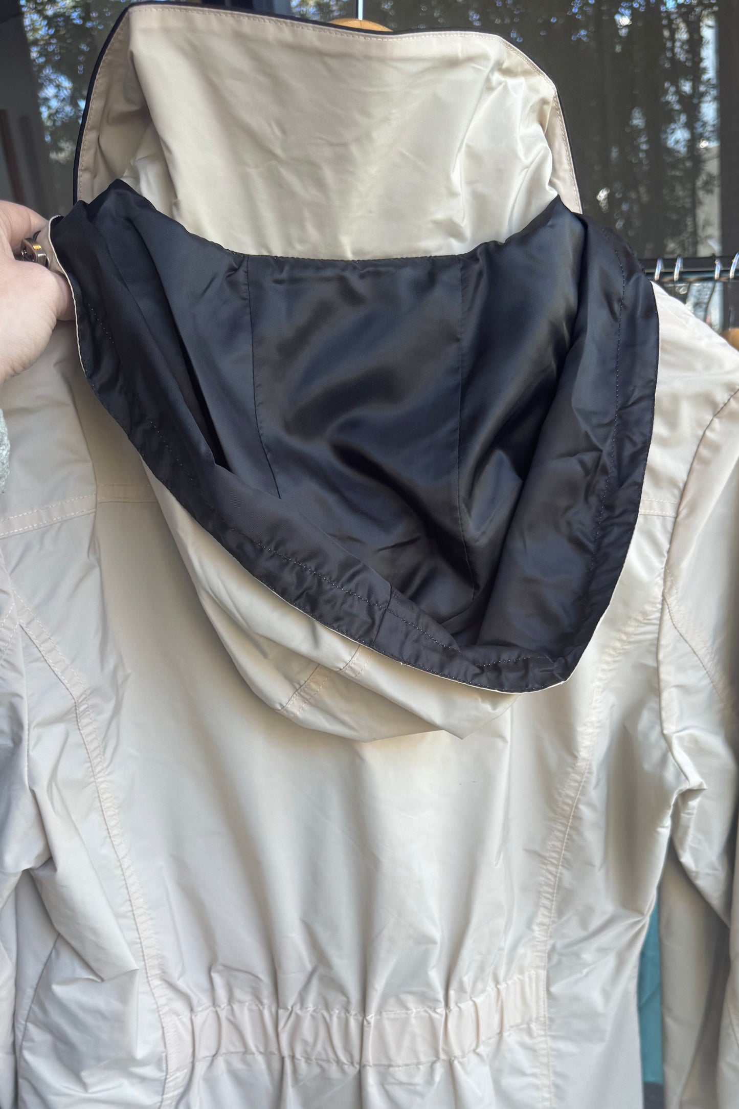 A cream-colored hooded jacket with a black inner hood lining, held up by a hand in front of a clothing store window.