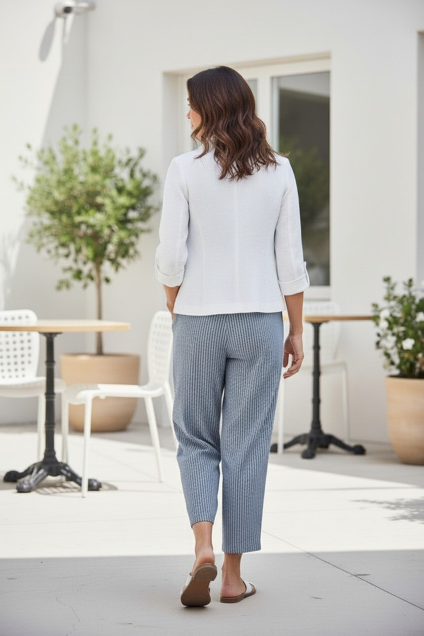 A woman viewed from behind wearing a white long-sleeve blouse and textured light blue cropped trousers, standing on a patio in front of a white building with outdoor tables and plants.