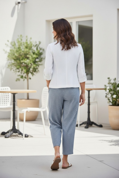 A woman viewed from behind wearing a white long-sleeve blouse and textured light blue cropped trousers, standing on a patio in front of a white building with outdoor tables and plants.