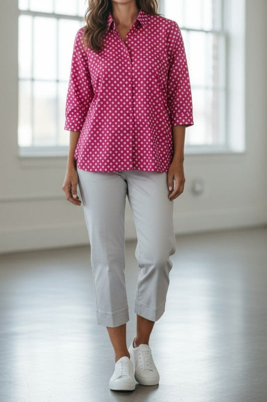 Woman wearing a pink polka dot shirt and white pants standing in a bright room with large windows.
